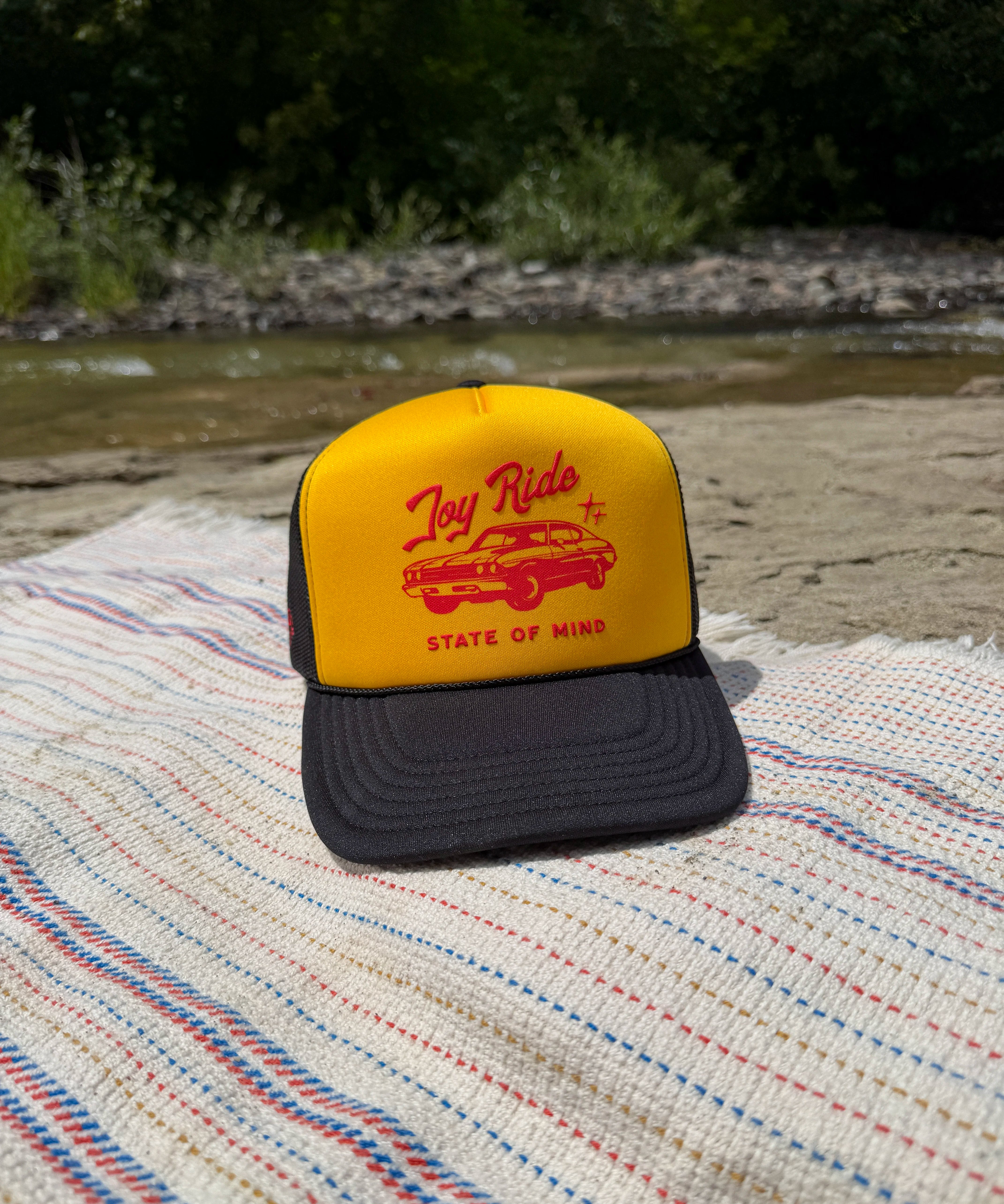 A yellow and black trucker hat with a red 'Joy Ride state of mind', resting on a striped blanket outdoors near a river and trees.