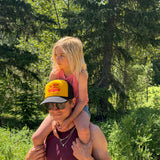 A man wearing a yellow and black trucker hat that says "joy ride state of mind". He carries a young blonde girl on his shoulders outdoors in a wooded area.