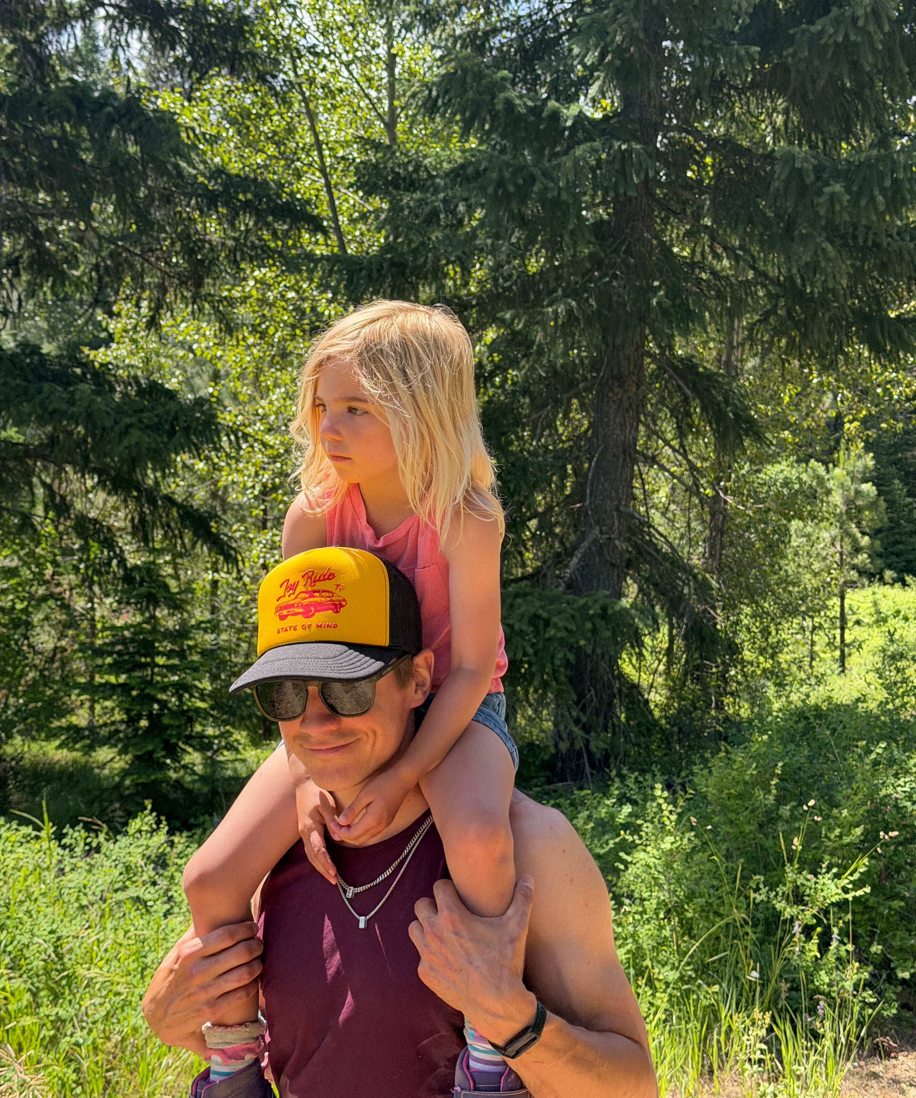 A man wearing a yellow and black trucker hat that says "joy ride state of mind". He carries a young blonde girl on his shoulders outdoors in a wooded area.