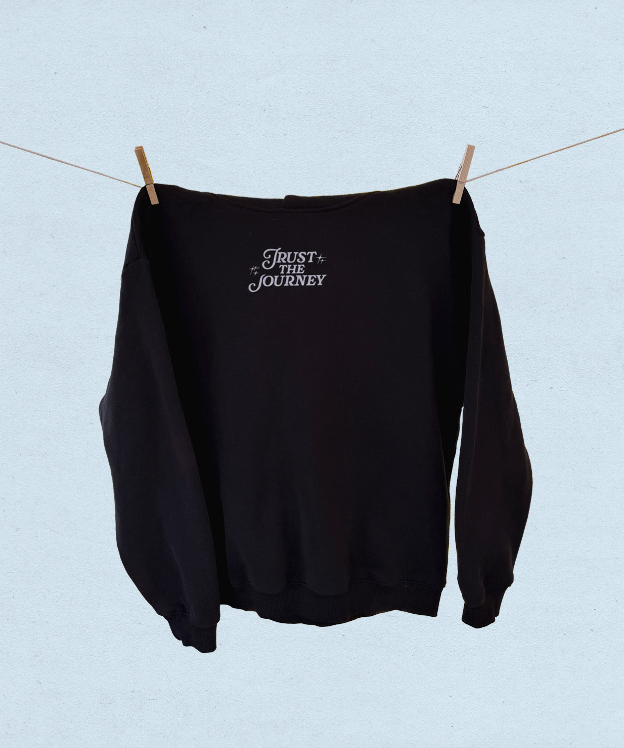 Black sweatshirt with 'Trust the Journey' text hanging on a clothesline against a light blue background