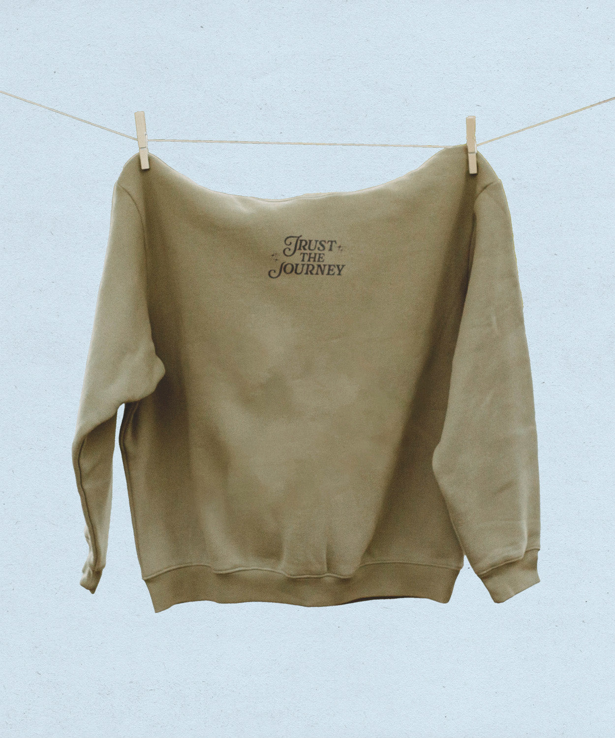 Front view of khaki hooded sweatshirt with 'Trust the Journey' text on a clothesline against a light blue background